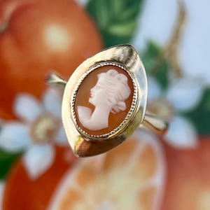 May include: A gold ring featuring an oval-shaped cameo with a light pink carved profile of a person. The cameo is set within a polished gold frame that has a pointed, marquise-like shape and a textured, beaded edge surrounding the central stone.
