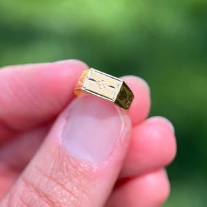 K&ouml;nnte beinhalten: Ein goldener Siegelring mit einer rechteckigen Platte, die ein eingraviertes Blumenmuster und einen kleinen zentralen Akzent aufweist. Die Ringschiene ist poliert und mit zus&auml;tzlichen dekorativen Gravuren an den Seiten versehen.