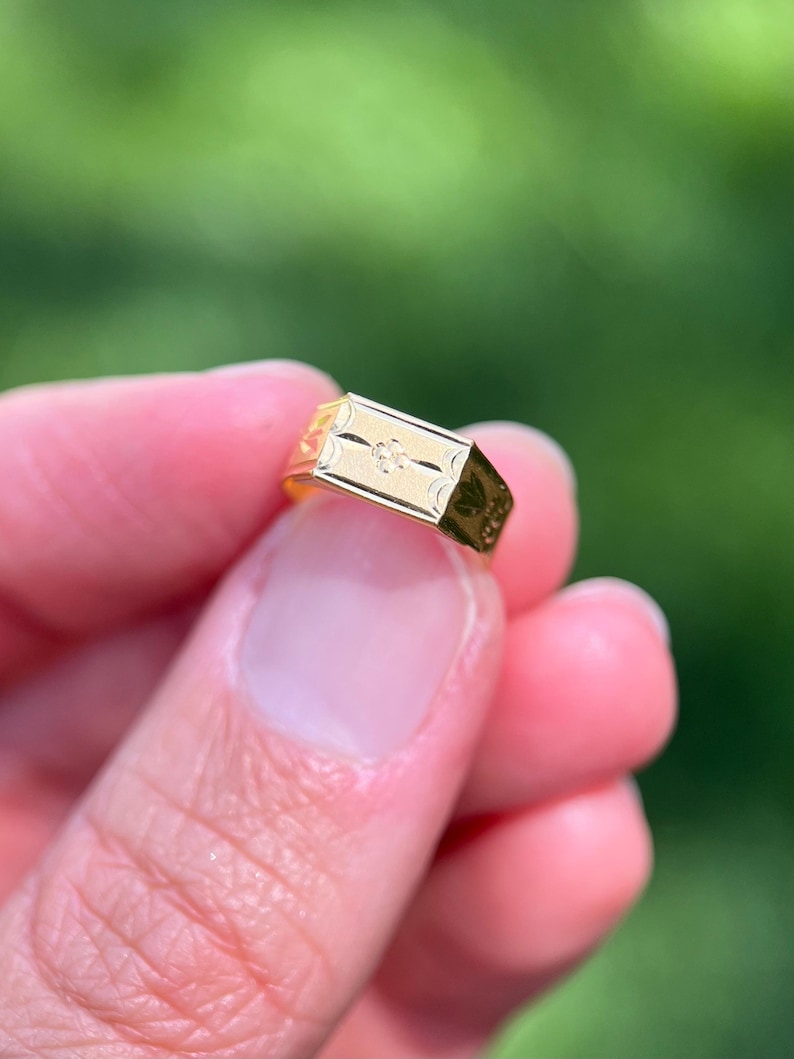 K&ouml;nnte beinhalten: Ein goldener Siegelring mit einer rechteckigen Platte, die ein eingraviertes Blumenmuster und einen kleinen zentralen Akzent aufweist. Die Ringschiene ist poliert und mit zus&auml;tzlichen dekorativen Gravuren an den Seiten versehen.