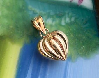 Könnte beinhalten: Ein goldfarbener Herzanhänger mit geripptem Design. Der Anhänger hat oben eine Schlaufe für eine Kette. Das Herz ist etwa 2,5 cm breit und 2,5 cm hoch. Der Hintergrund ist eine Mischung aus Blau und Grün.