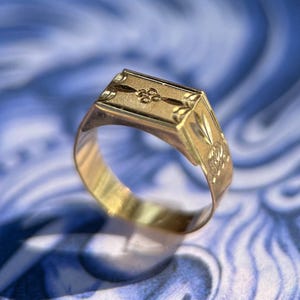 K&ouml;nnte beinhalten: Ein goldener Siegelring mit rechteckiger Platte, die ein eingraviertes zentrales Blumenmotiv und dekorative &Auml;tzmuster an den Seiten aufweist. Die Ringschiene ist glatt und poliert und zeigt durchgehend ein metallisch gelbes Finish.
