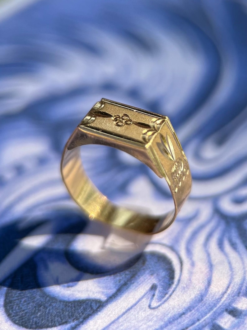 K&ouml;nnte beinhalten: Ein goldener Siegelring mit rechteckiger Platte, die ein eingraviertes zentrales Blumenmotiv und dekorative &Auml;tzmuster an den Seiten aufweist. Die Ringschiene ist glatt und poliert und zeigt durchgehend ein metallisch gelbes Finish.