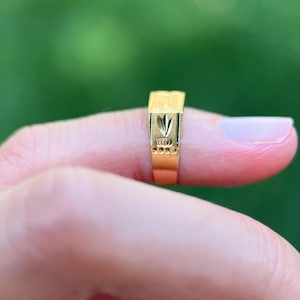 K&ouml;nnte beinhalten: Ein goldener Ring mit einer rechteckigen Oberseite, die ein eingraviertes blattartiges Motiv aufweist, sowie einer Reihe kleiner kreisf&ouml;rmiger Vertiefungen entlang der Seite der Ringschiene. Das Metall hat eine polierte Oberfl&auml;che.