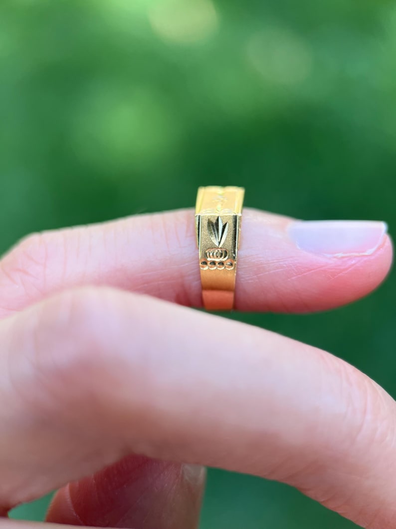 K&ouml;nnte beinhalten: Ein goldener Ring mit einer rechteckigen Oberseite, die ein eingraviertes blattartiges Motiv aufweist, sowie einer Reihe kleiner kreisf&ouml;rmiger Vertiefungen entlang der Seite der Ringschiene. Das Metall hat eine polierte Oberfl&auml;che.