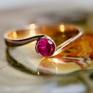 Puede incluir: Un anillo de oro con una sola piedra preciosa de rubí rosa oscuro redonda engastada en el centro. La banda es curva y tiene un acabado liso y pulido.