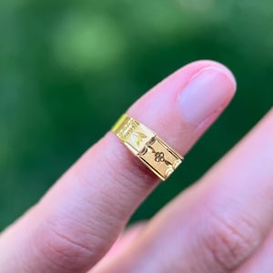 K&ouml;nnte beinhalten: Ein goldener Bandring mit einer Reihe sich wiederholender rechteckiger Paneele. Jedes Paneel ist mit einem floralen Motiv graviert, darunter eine kleine vierbl&auml;ttrige Blume und ein Blattdesign, auf einer glatten, polierten Goldoberfl&auml;che.