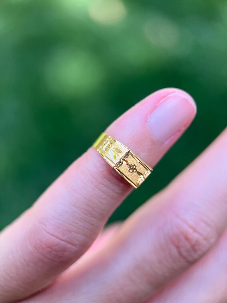 K&ouml;nnte beinhalten: Ein goldener Bandring mit einer Reihe sich wiederholender rechteckiger Paneele. Jedes Paneel ist mit einem floralen Motiv graviert, darunter eine kleine vierbl&auml;ttrige Blume und ein Blattdesign, auf einer glatten, polierten Goldoberfl&auml;che.