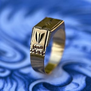 K&ouml;nnte beinhalten: Ein goldener Siegelring mit einer rechteckigen, flachen Oberseite und einem eingravierten Blumenmotiv. Die Seite des Rings zeigt ein vertikales blattartiges Muster &uuml;ber einer Reihe kleiner kreisf&ouml;rmiger Vertiefungen mit polierter Oberfl&auml;che.