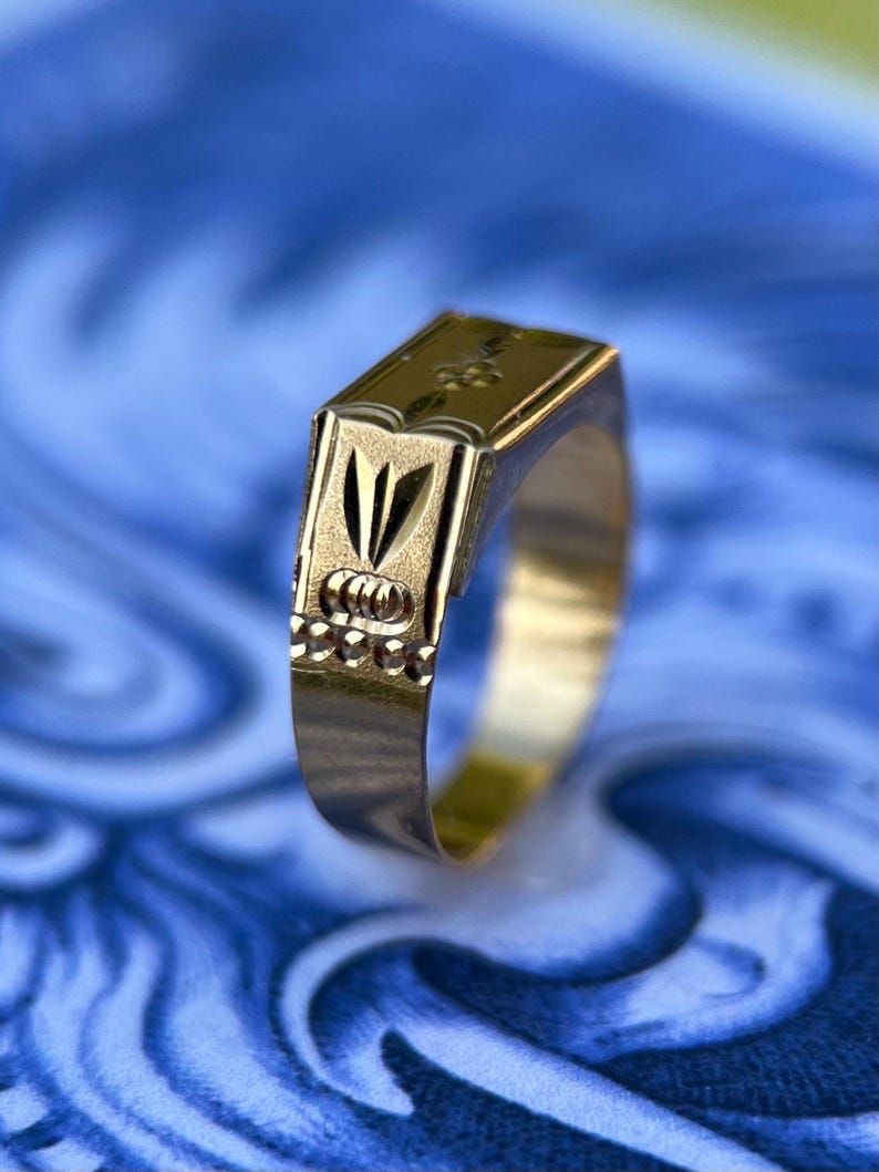 K&ouml;nnte beinhalten: Ein goldener Siegelring mit einer rechteckigen, flachen Oberseite und einem eingravierten Blumenmotiv. Die Seite des Rings zeigt ein vertikales blattartiges Muster &uuml;ber einer Reihe kleiner kreisf&ouml;rmiger Vertiefungen mit polierter Oberfl&auml;che.