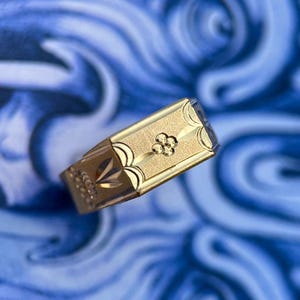 K&ouml;nnte beinhalten: Ein goldener Siegelring mit einer rechteckigen Platte in matter, texturierter Ausf&uuml;hrung. In der Mitte der Platte befindet sich ein kleines, vierbl&auml;ttriges Blumenmotiv. Die Seiten des Rings sind mit gravierten Bogenlinien und Mustern verziert.