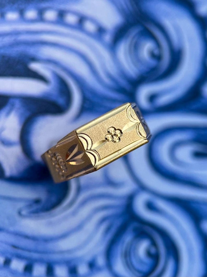 K&ouml;nnte beinhalten: Ein goldener Siegelring mit einer rechteckigen Platte in matter, texturierter Ausf&uuml;hrung. In der Mitte der Platte befindet sich ein kleines, vierbl&auml;ttriges Blumenmotiv. Die Seiten des Rings sind mit gravierten Bogenlinien und Mustern verziert.