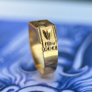 K&ouml;nnte beinhalten: Ein Goldring mit einer flachen, rechteckigen Oberseite, die ein eingraviertes Design aus einem stilisierten Blattmotiv &uuml;ber einer Reihe von drei horizontalen, ovalen Aussparungen aufweist, eingefasst in eine polierte Goldschiene.