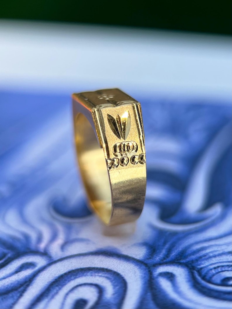 K&ouml;nnte beinhalten: Ein Goldring mit einer flachen, rechteckigen Oberseite, die ein eingraviertes Design aus einem stilisierten Blattmotiv &uuml;ber einer Reihe von drei horizontalen, ovalen Aussparungen aufweist, eingefasst in eine polierte Goldschiene.
