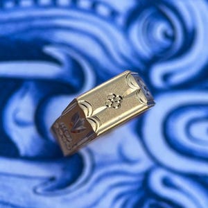K&ouml;nnte beinhalten: Ein goldfarbener, rechteckiger Siegelring mit einer zentralen, vierbl&auml;ttrigen Blumengravur auf matter Oberfl&auml;che, flankiert von geschwungenen Linien an den Seiten und weiteren dekorativen Mustern entlang der Ringschiene.