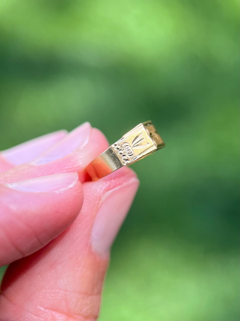 K&ouml;nnte beinhalten: Ein goldener Siegelring mit einer rechteckigen Platte, die ein eingraviertes Blumenmotiv aufweist. Die Ringschiene ist glatt und poliert und hat eine metallisch gelbe Oberfl&auml;che. Das Design enth&auml;lt ein kleines, detailliertes florales Muster.