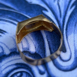 K&ouml;nnte beinhalten: Ein goldener Ring mit einer flachen, rechteckigen Oberseite, der eine Gravur oder einen Stempel auf der Innenseite des Rings aufweist. Der Ring hat eine polierte metallische Oberfl&auml;che und ein massives, durchgehendes Band, das sich nach oben hin leicht verj&uuml;ngt.