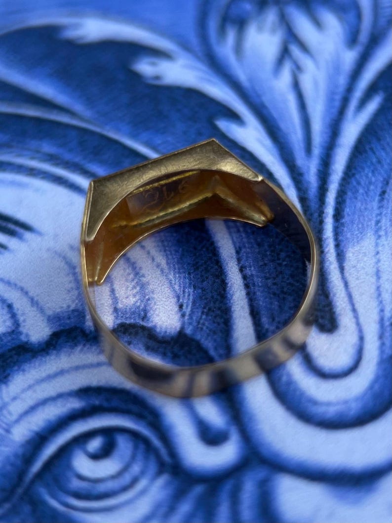 K&ouml;nnte beinhalten: Ein goldener Ring mit einer flachen, rechteckigen Oberseite, der eine Gravur oder einen Stempel auf der Innenseite des Rings aufweist. Der Ring hat eine polierte metallische Oberfl&auml;che und ein massives, durchgehendes Band, das sich nach oben hin leicht verj&uuml;ngt.