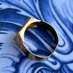 K&ouml;nnte beinhalten: Ein goldfarbener Metallring mit einer flachen, kantigen Oberseite und einer glatten, kreisf&ouml;rmigen Ringschiene. Der Ring hat eine polierte Oberfl&auml;che, die das Licht reflektiert und ein schlichtes, geometrisches Design mit klaren Linien zeigt.