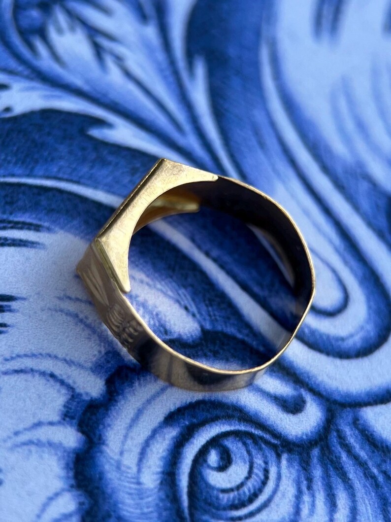 K&ouml;nnte beinhalten: Ein goldfarbener Metallring mit einer flachen, kantigen Oberseite und einer glatten, kreisf&ouml;rmigen Ringschiene. Der Ring hat eine polierte Oberfl&auml;che, die das Licht reflektiert und ein schlichtes, geometrisches Design mit klaren Linien zeigt.