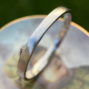 May include: A silver-toned metal hinged bangle bracelet with a brushed, textured finish. The bracelet features a secure clasp mechanism and a thin safety chain attached to the side, showcasing a clean, rounded profile and a polished inner band.