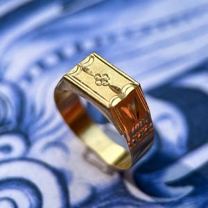 K&ouml;nnte beinhalten: Ein goldener Siegelring mit rechteckiger Platte, die ein eingraviertes vierbl&auml;ttriges Blumenmotiv in der Mitte einer horizontalen Linie aufweist. Die Seiten des Rings zeigen ge&auml;tzte geometrische Muster und die Ringschiene ist poliert.