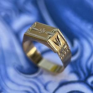 K&ouml;nnte beinhalten: Ein Goldring mit einer rechteckigen Kopfplatte, die ein eingraviertes Blumenmotiv aufweist, sowie einem texturierten Seitenteil mit f&auml;cherf&ouml;rmigem Design. Die Ringschiene ist glatt und poliert und zeigt eine Kombination aus matten und gl&auml;nzenden Goldoberfl&auml;chen.