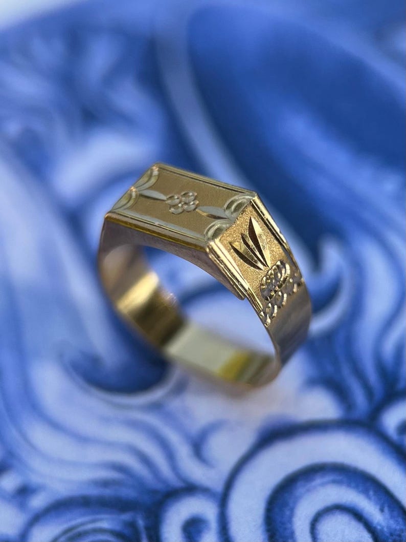 K&ouml;nnte beinhalten: Ein Goldring mit einer rechteckigen Kopfplatte, die ein eingraviertes Blumenmotiv aufweist, sowie einem texturierten Seitenteil mit f&auml;cherf&ouml;rmigem Design. Die Ringschiene ist glatt und poliert und zeigt eine Kombination aus matten und gl&auml;nzenden Goldoberfl&auml;chen.