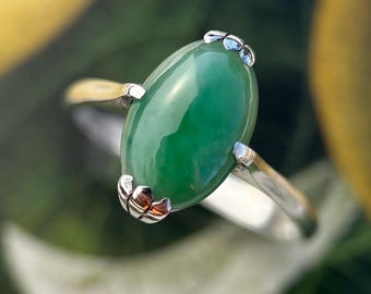 Könnte beinhalten: Nahaufnahme eines Rings mit einem großen, ovalen, leuchtend grünen Edelstein. Der Stein ist in einem silbernen Band mit dekorativen Akzenten gefasst. Der Ring ist vor einem verschwommenen grünen und gelben Hintergrund zu sehen.