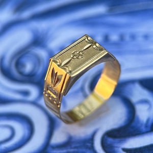 K&ouml;nnte beinhalten: Ein goldener Siegelring mit rechteckiger Platte, die ein eingraviertes Blumenmotiv sowie dekorative geometrische Muster an den Seiten aufweist. Die Ringschiene ist poliert und konisch zulaufend gestaltet und zeigt feine Metallverzierungen.