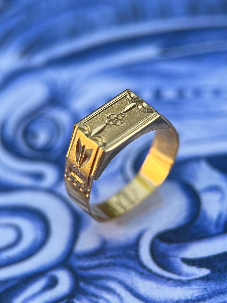 K&ouml;nnte beinhalten: Ein goldener Siegelring mit rechteckiger Platte, die ein eingraviertes Blumenmotiv sowie dekorative geometrische Muster an den Seiten aufweist. Die Ringschiene ist poliert und konisch zulaufend gestaltet und zeigt feine Metallverzierungen.