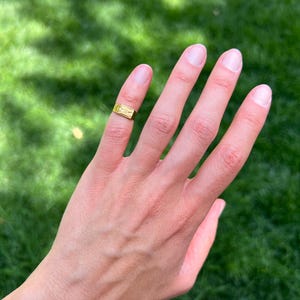K&ouml;nnte beinhalten: Ein goldfarbener Metallring mit einer rechteckigen Vorderseite und einer eingravierten Verzierung. Der Ring wird am Mittelfinger getragen und zeichnet sich durch eine polierte Oberfl&auml;che sowie eine flache, horizontale Ringschiene aus.