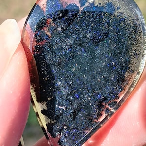 May include: A teardrop-shaped pendant filled with dark blue glitter and resin. The clear resin allows the glitter to sparkle. The edges of the pendant have a copper-colored shimmer. A small hole is at the top for a chain.