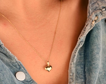 May include: A gold heart pendant necklace on a chain. The necklace is worn by a person wearing a denim shirt.