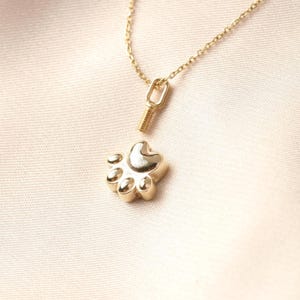 May include: A gold-colored paw print pendant necklace. The pendant is a detailed paw print with five pads and a heart-shaped center. The necklace chain and clasp are also gold-colored. The necklace is displayed on a light pink fabric.