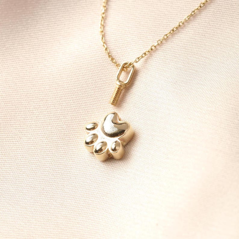 May include: A gold-colored paw print pendant necklace. The pendant is a detailed paw print with five pads and a heart-shaped center. The necklace chain and clasp are also gold-colored. The necklace is displayed on a light pink fabric.