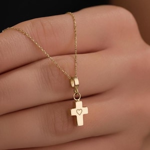 Può includere: Una delicata collana color oro con un piccolo pendente a forma di croce con un cuore. La croce pende da una sottile catena, creando un design semplice ma elegante. La collana è mostrata su uno sfondo neutro.