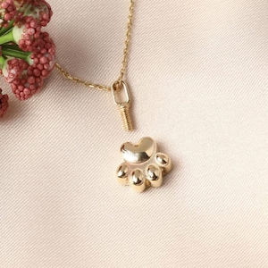 May include: A gold-colored necklace featuring a paw print pendant. The pendant has five rounded toe pads and a heart-shaped top pad. The necklace chain and clasp are also gold-colored. The background is a light, neutral color.