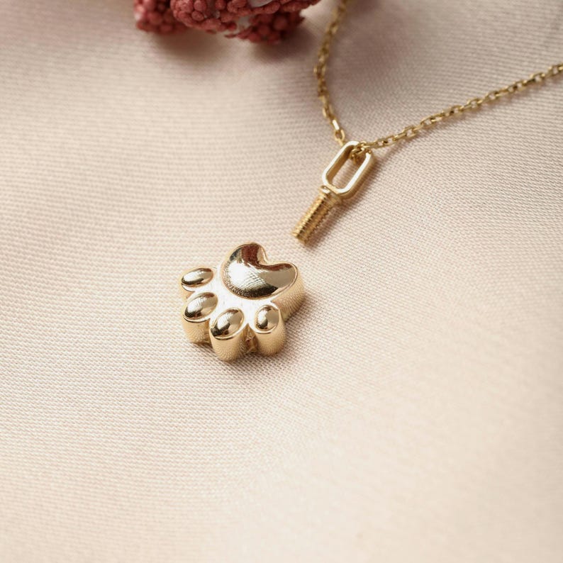 May include: A gold-colored paw print pendant with a matching chain. The pendant is detailed with five rounded pads and a heart-shaped top. The chain is delicate, and the pendant is detached from the clasp. The background is a soft, neutral color.
