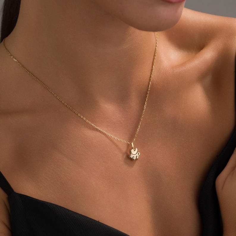 May include: A delicate gold necklace with a small pendant. The pendant has a detailed, ornate design. The necklace is worn against a dark top, creating a contrast that highlights the jewelry. The chain is thin and elegant.