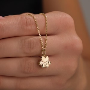 May include: A gold-colored necklace featuring a paw print pendant. The pendant has a heart-shaped top and five rounded pads. The chain is delicate and the necklace is held in a person's hand.