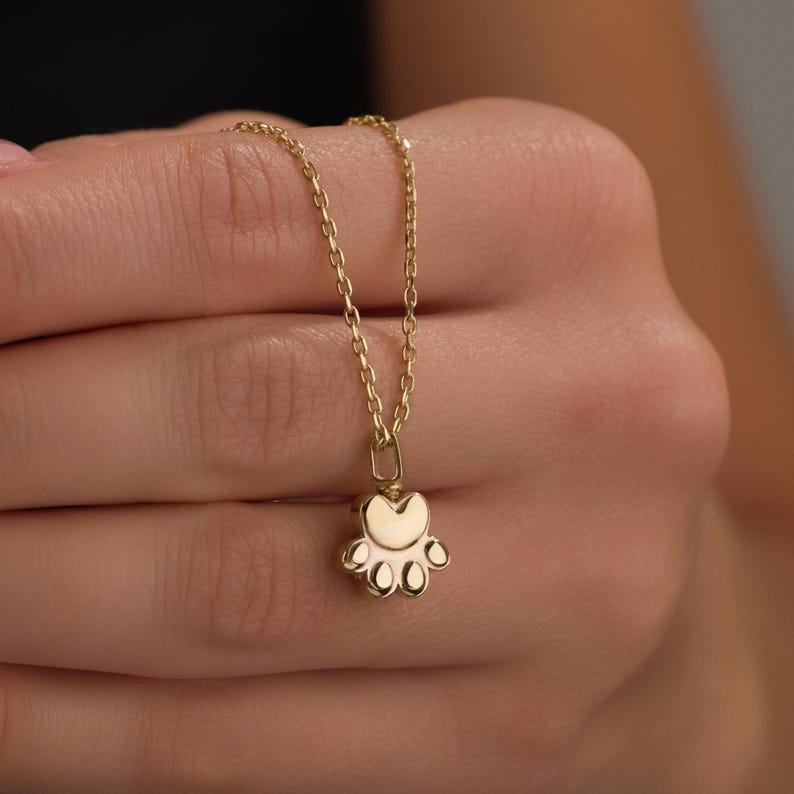 May include: A gold-colored necklace featuring a paw print pendant. The pendant has a heart-shaped top and five rounded pads. The chain is delicate and the necklace is held in a person's hand.