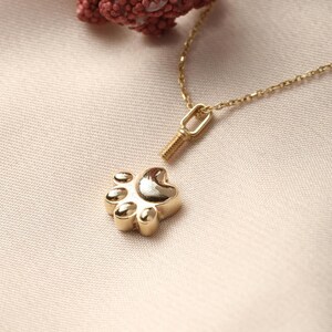 May include: A gold-colored necklace featuring a paw print pendant. The pendant has a detailed paw print design with rounded pads. The necklace chain is delicate and gold, with a small clasp. The background is a soft, neutral color.