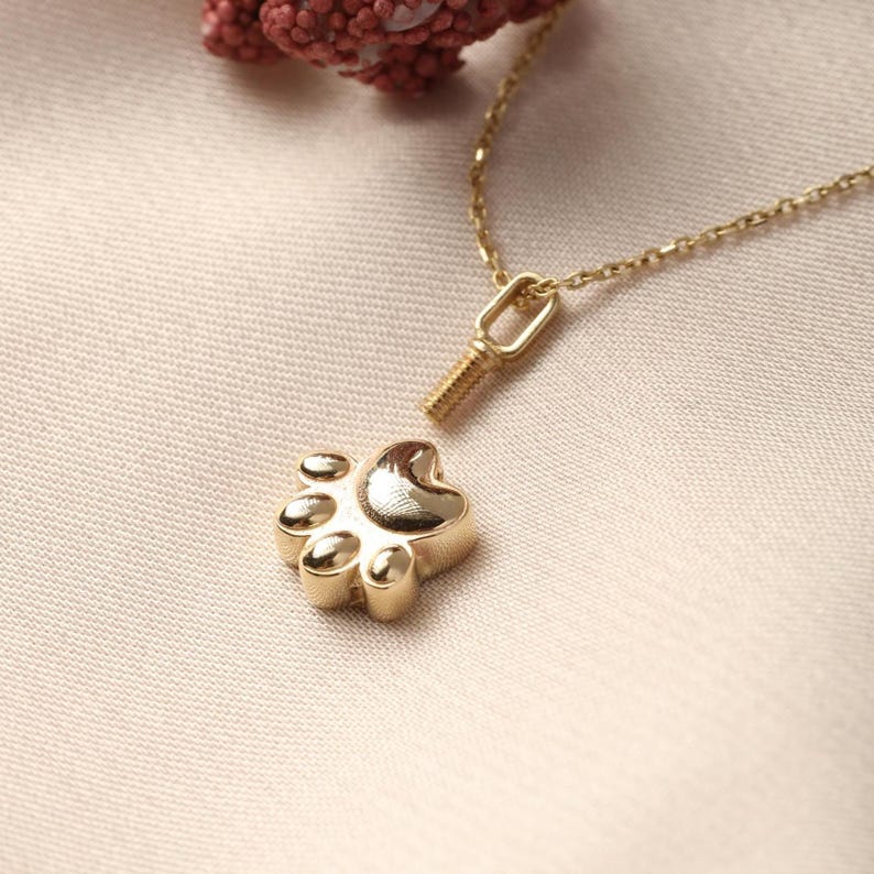 May include: A gold-colored necklace featuring a paw print pendant. The pendant has a detailed paw print design with rounded pads. The necklace chain is delicate and gold, with a small clasp. The background is a soft, neutral color.