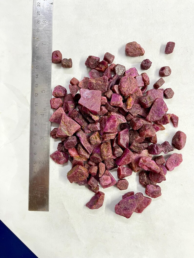 Op de afbeelding: Een verzameling ruwe, ongeslepen robijn edelstenen in verschillende maten en vormen. De edelstenen hebben een dieprode kleur. Een metalen liniaal met centimeter maten ligt ernaast.