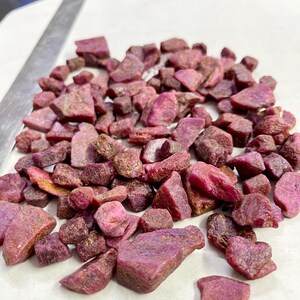 Op de afbeelding: Een verzameling ruwe, ongeslepen robijn edelstenen in verschillende maten en vormen. De edelstenen vertonen een dieprode tot paarse tint, met enkele lichtere tinten en een paar gouden insluitsels. De robijnen zijn verspreid over een wit oppervlak.