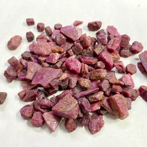 Op de afbeelding: Een stapel ruwe robijn edelstenen in verschillende maten en vormen. De robijnen vertonen een reeks dieprode en roze tinten, waarbij sommige een bruinachtige tint vertonen. De edelstenen zijn verspreid over een wit oppervlak.