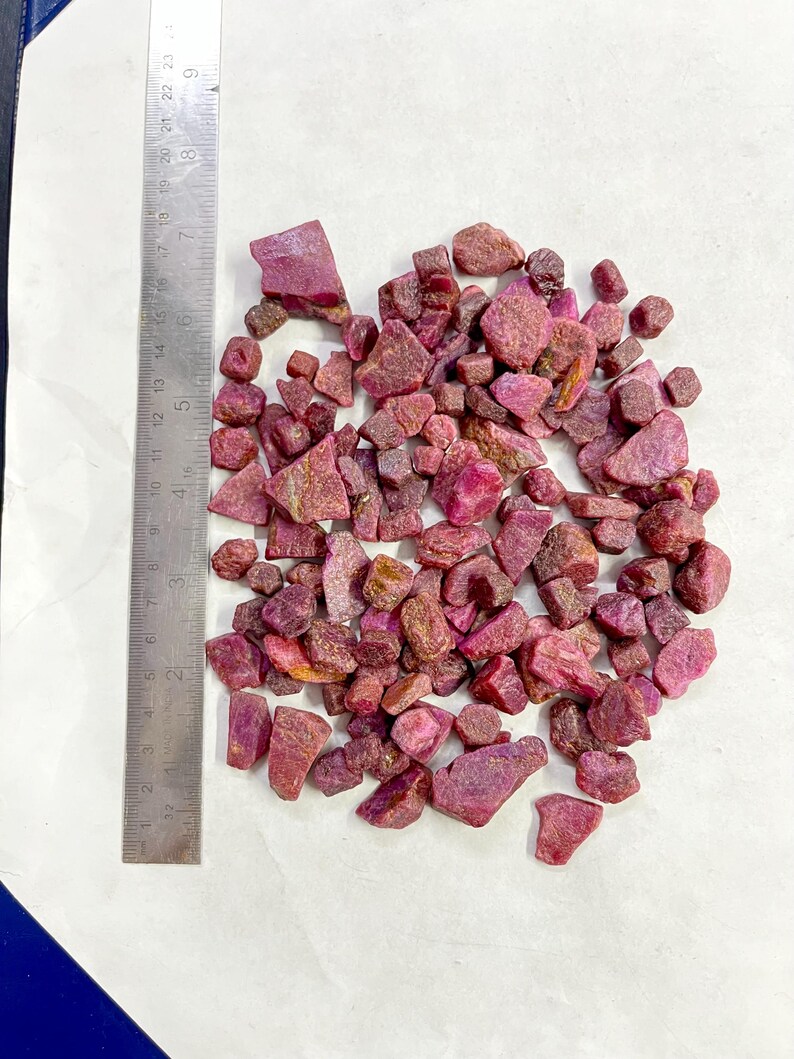 Op de afbeelding: Een verzameling ruwe, ongeslepen robijn edelstenen in verschillende tinten rood en roze. De edelstenen zijn verspreid op een wit oppervlak naast een metalen liniaal.