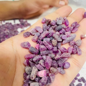 May include: A handful of rough, uncut ruby gemstones in shades of pink and purple. The gemstones are irregularly shaped and vary in size. The image shows a close-up view of the gemstones, highlighting their natural texture and color.