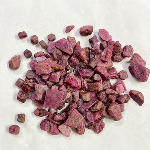 Op de afbeelding: Een stapel ruwe robijn edelstenen in verschillende vormen en maten. De edelstenen vertonen een reeks dieprode en roze tinten, waarbij sommige een paarsachtige tint vertonen. De ruwe stenen liggen verspreid op een wit oppervlak.