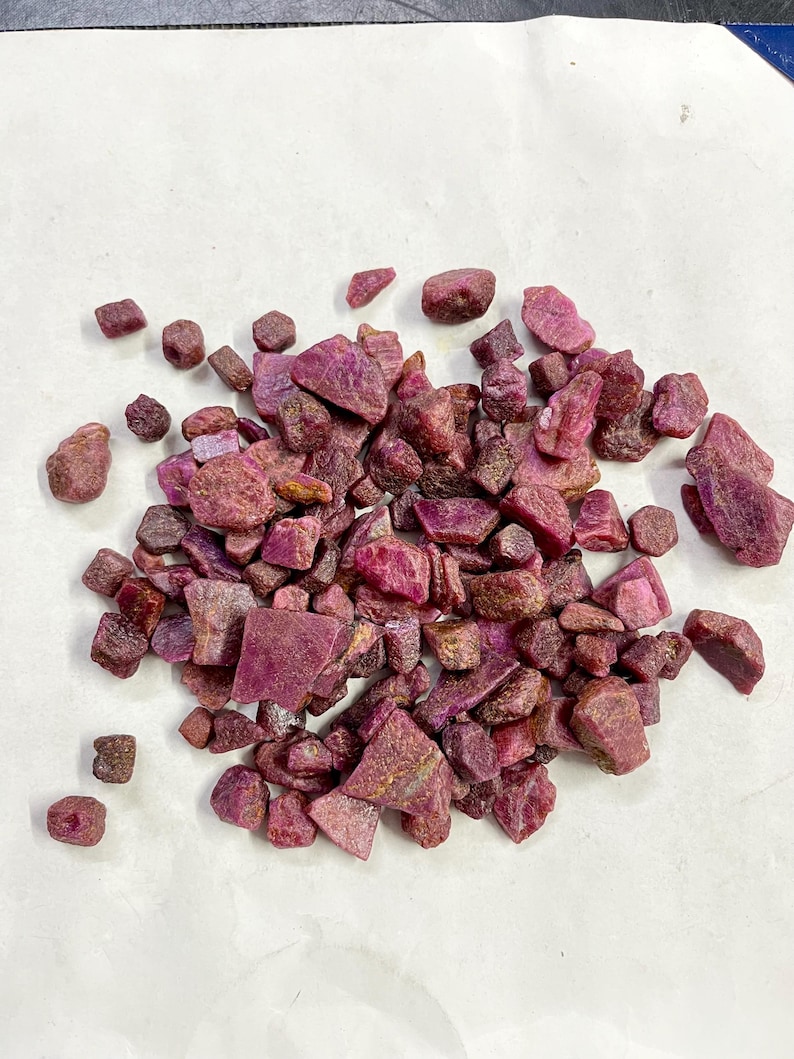 Op de afbeelding: Een stapel ruwe robijn edelstenen in verschillende vormen en maten. De edelstenen vertonen een reeks dieprode en roze tinten, waarbij sommige een paarsachtige tint vertonen. De ruwe stenen liggen verspreid op een wit oppervlak.